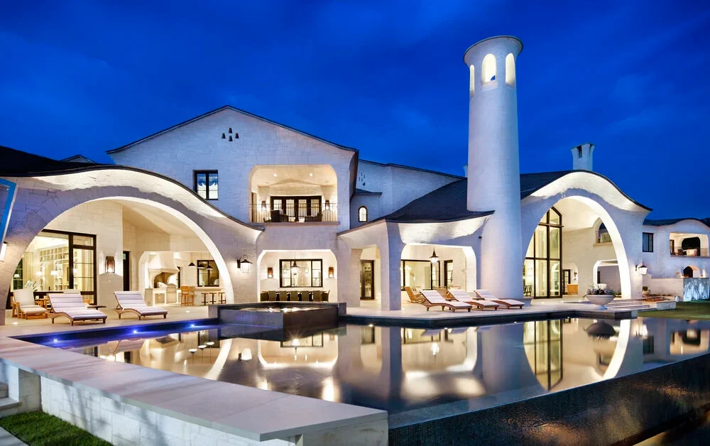 Curved white-stone estate with arched loggias glowing at dusk, mirrored in a dark geometric pool