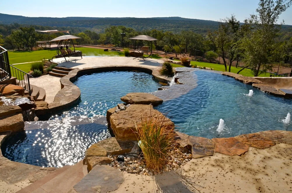 Freeform pool with stone spillways and a raised spa overlooking a Hill Country lawn and distant ridgelines