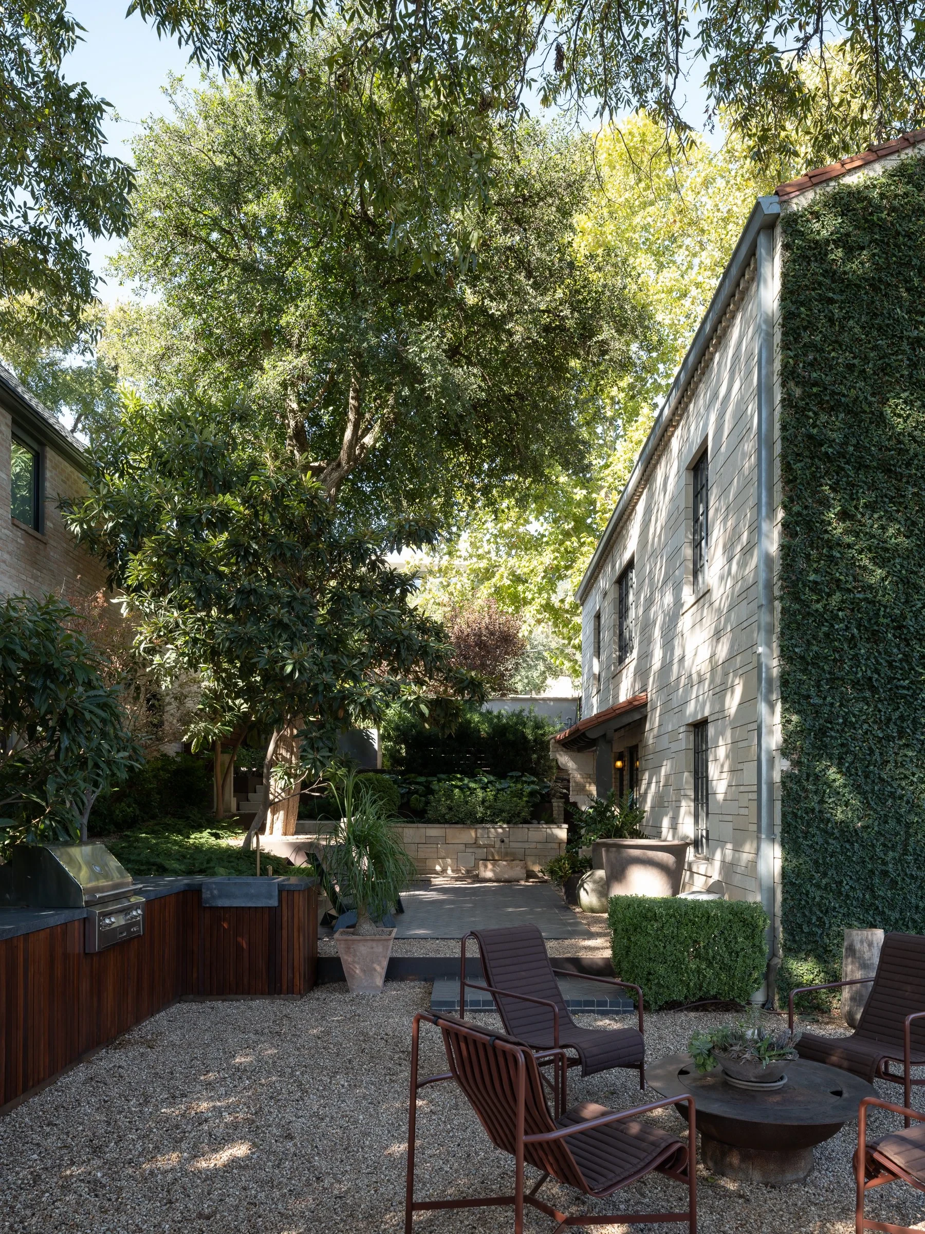 Gravel courtyard with red metal chairs and a wood bar under a tall tree beside an ivy-clad home