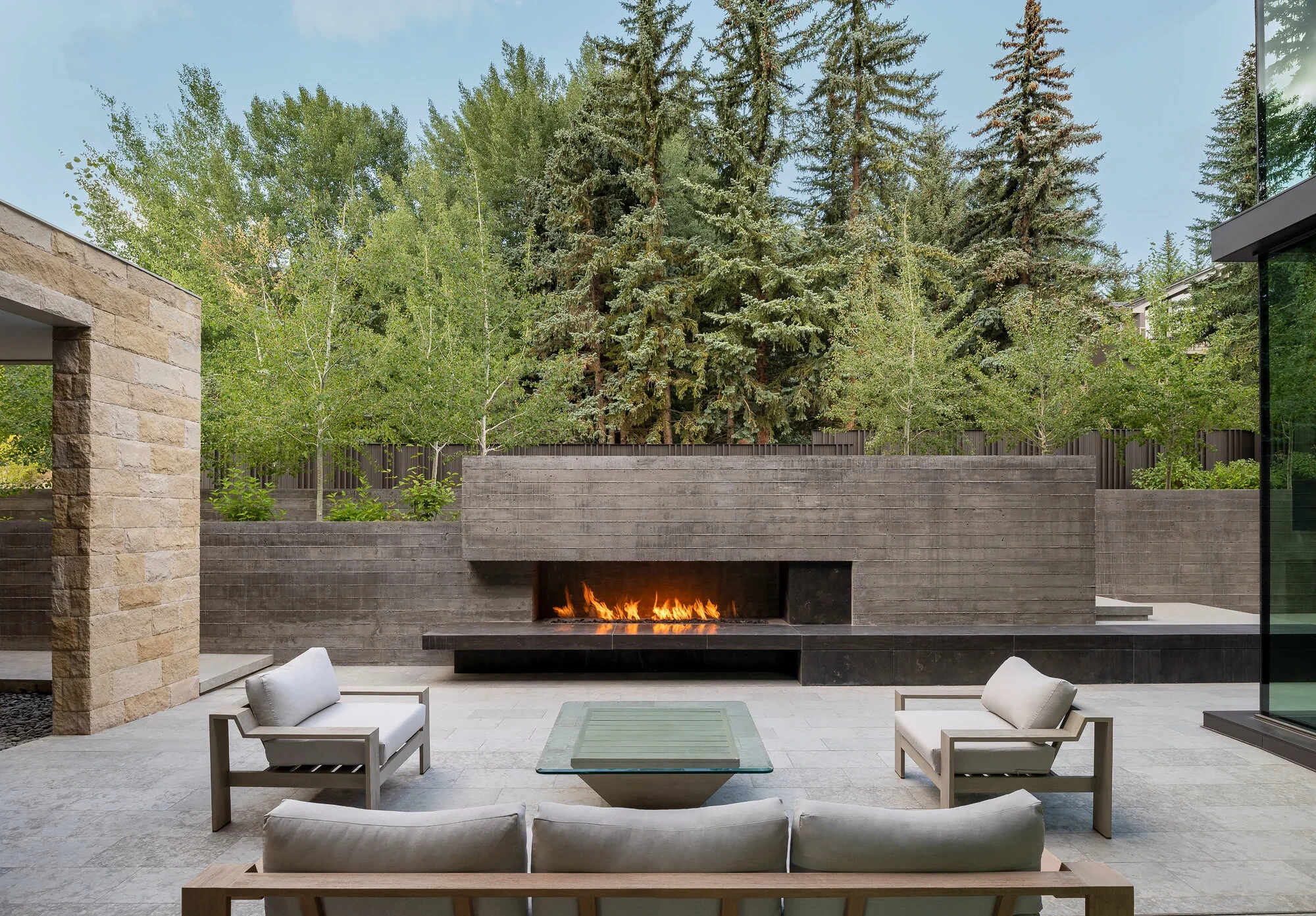 Stone courtyard with a long linear gas fireplace, wood lounge seating, and tall evergreens behind