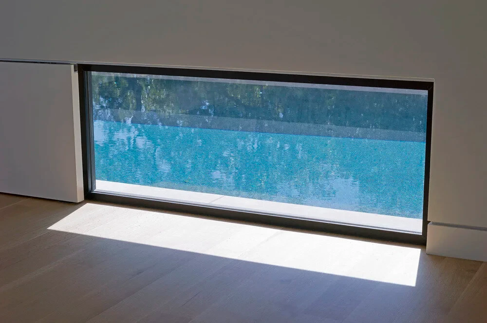 Pale wood floor and a low picture window framing the turquoise water of an infinity pool outside