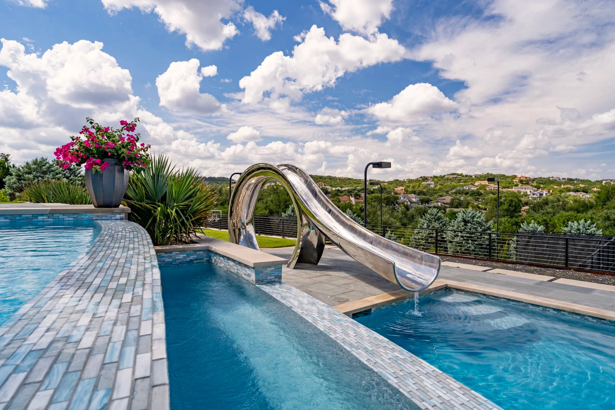Lakeway pool with a polished stainless waterslide spilling into blue tile, hilltop neighborhood view beyond