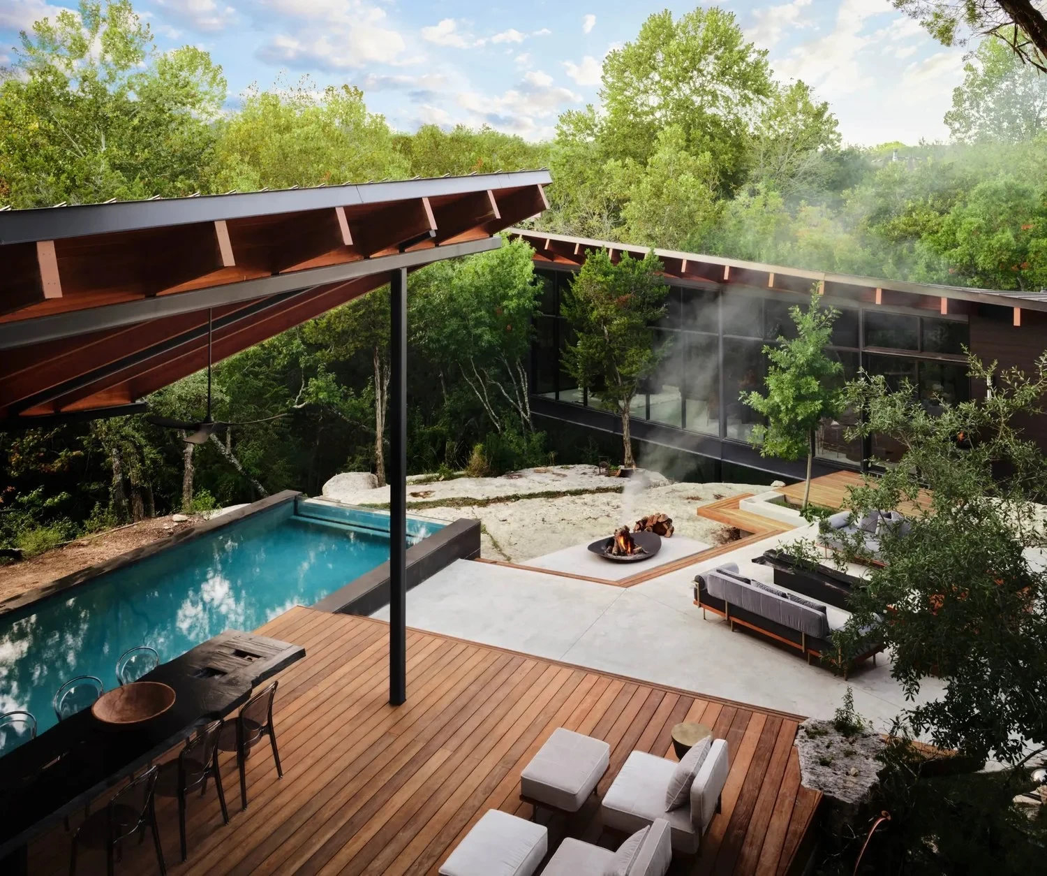 Geometric pool and spa beside a steel-and-wood glass pavilion, with an ipe deck, fire pit lounge and dense woods