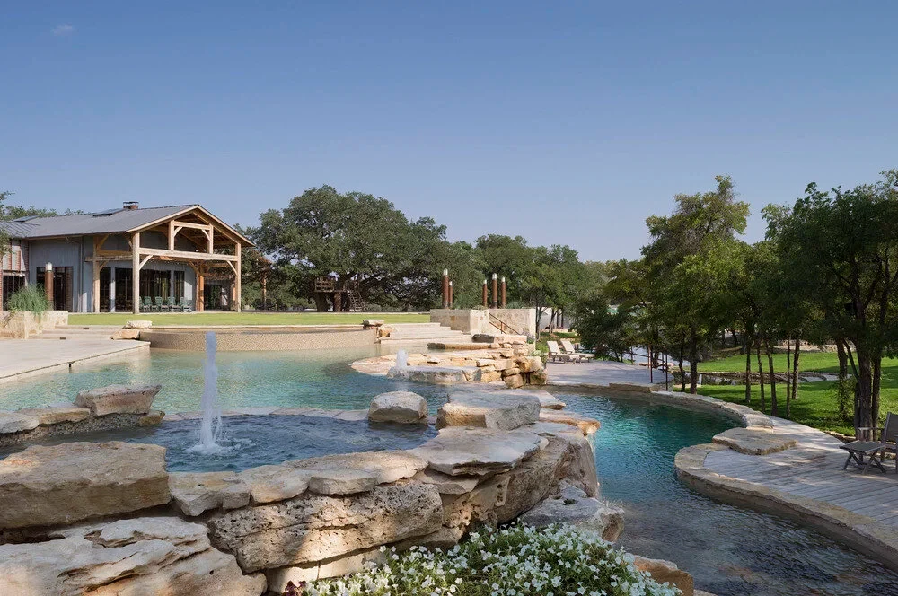 Freeform pool with rock waterfalls and a tanning ledge below a timber-framed lodge, ringed by oaks