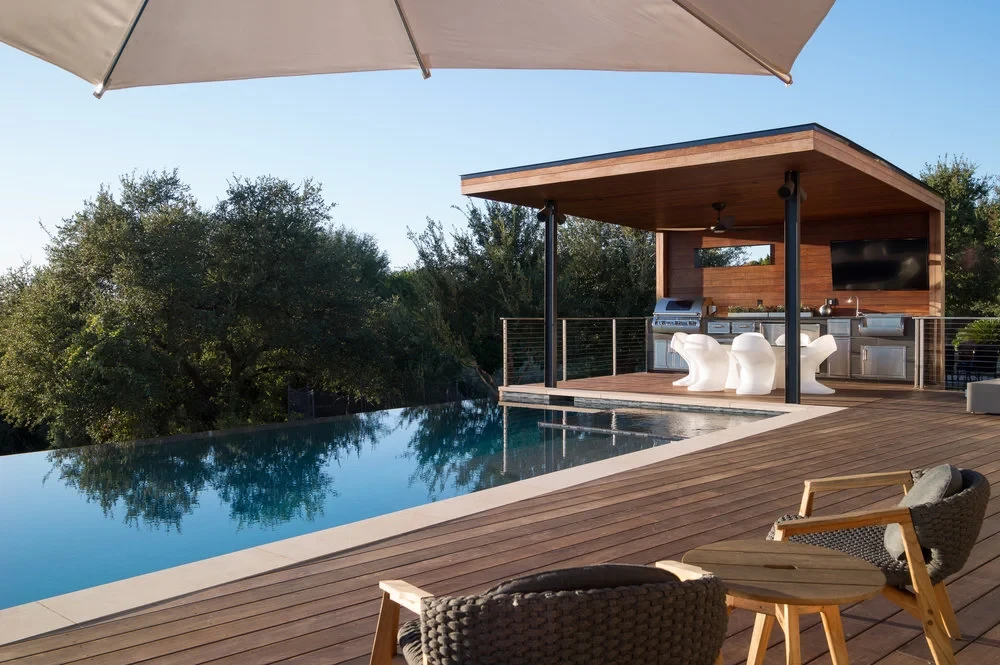Hill Country infinity pool with a wood deck, poolside cabana and outdoor kitchen under a shade umbrella