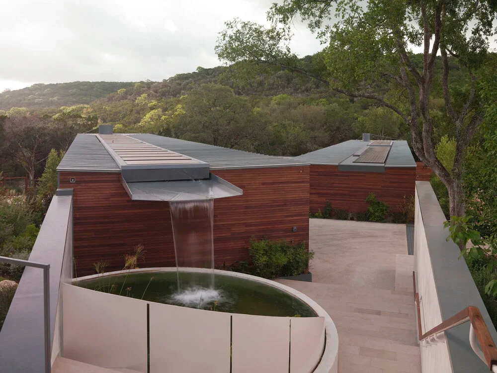 Round stone water-feature pool with a sheer waterfall off a wood-clad roof, Hill Country hills beyond