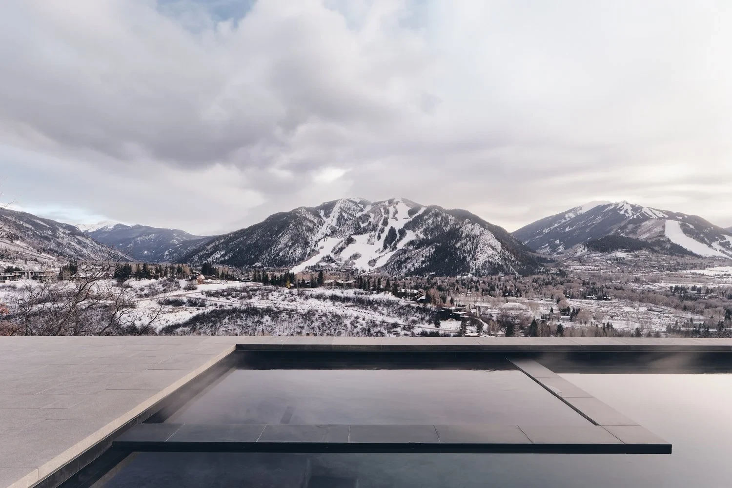 Dark infinity pool edge framing snow-covered Aspen mountains under a gray winter sky