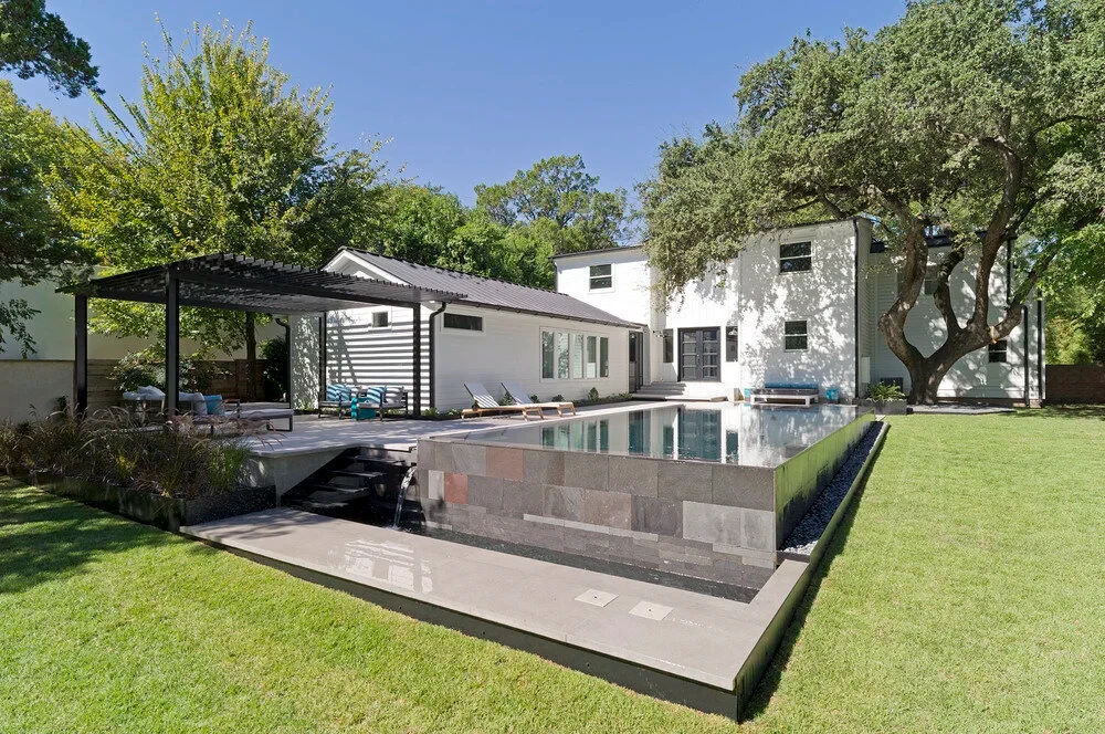 Dark geometric pool with a perimeter-overflow edge beside a steel pergola and green lawn at a modern home