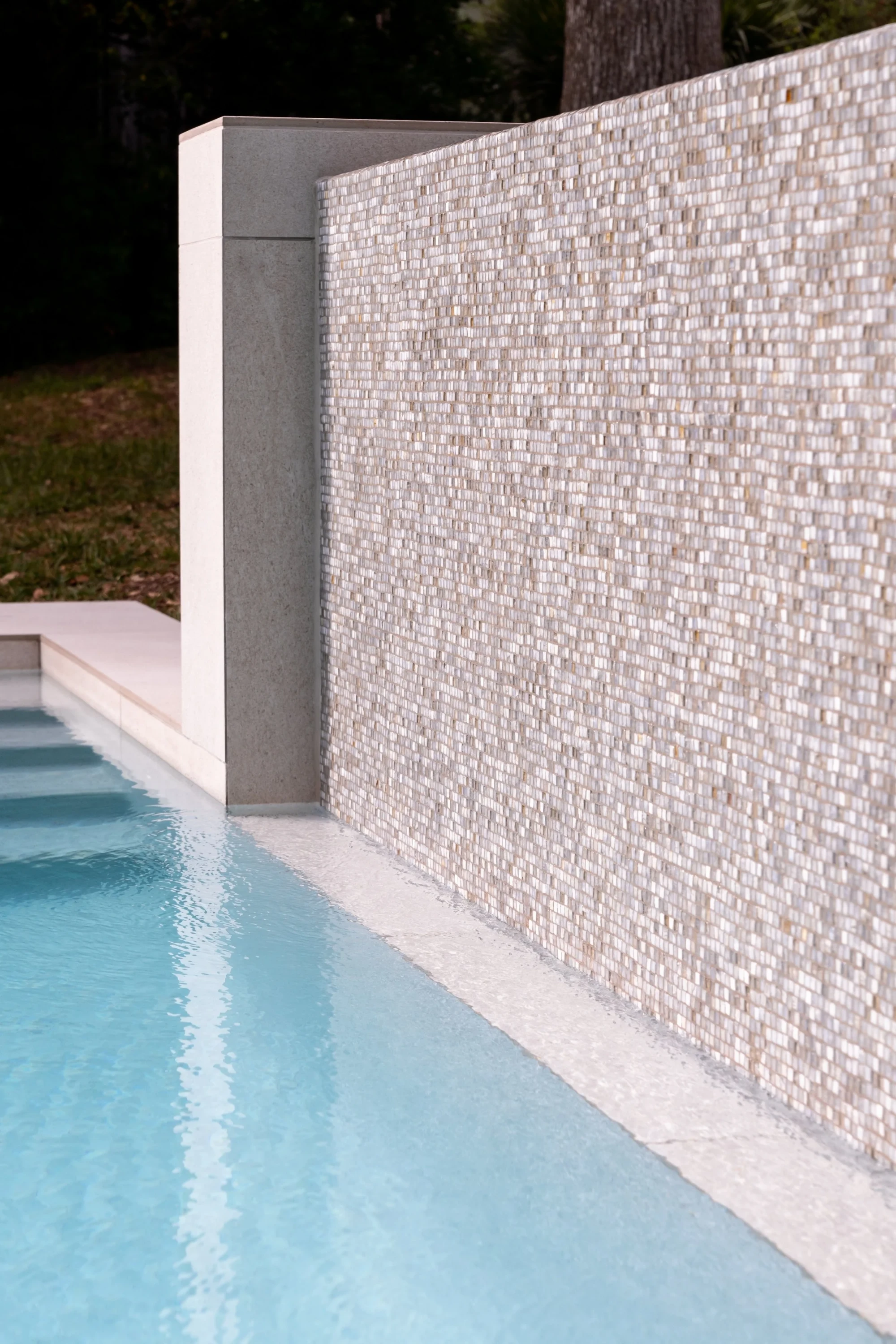 Pool edge with a pebbled stone water wall and limestone coping, wooded West Austin yard beyond