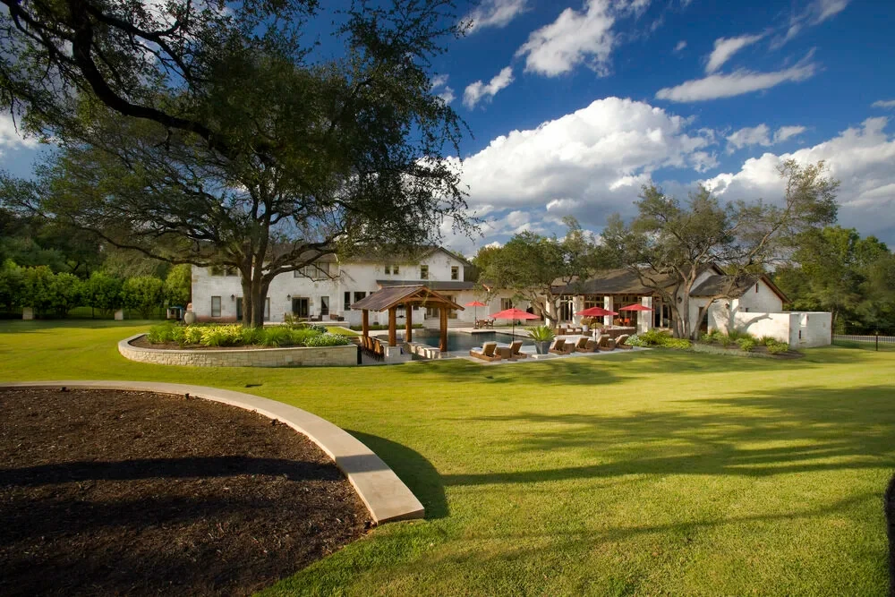 Stone estate behind a wide green lawn and oaks with a pool and red umbrellas in West Lake Hills