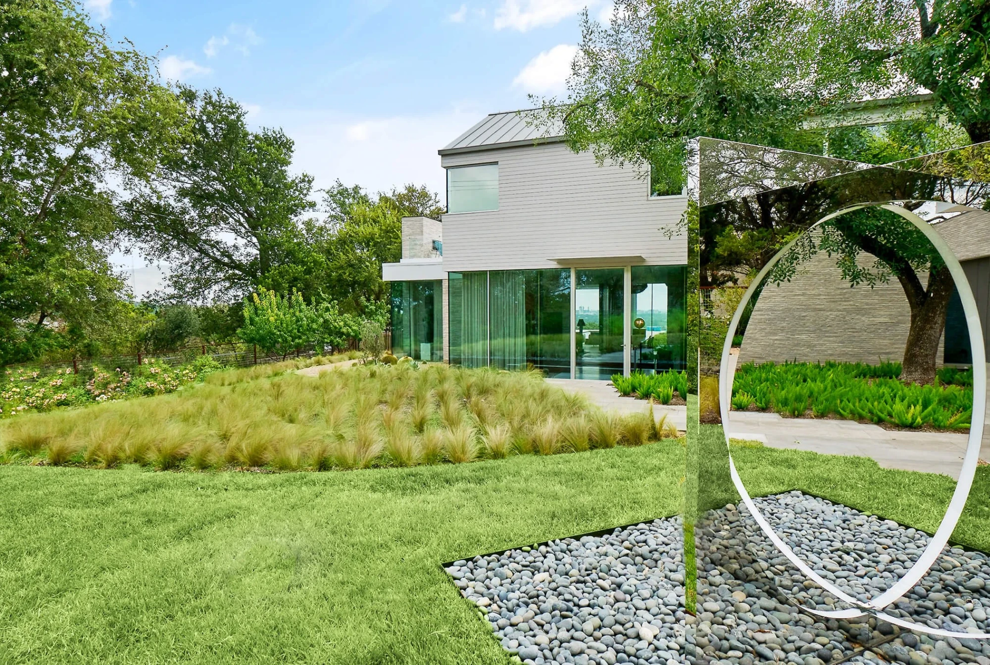Modern gray home with a metal roof above ornamental grasses, a river-rock bed and a tall circular sculpture