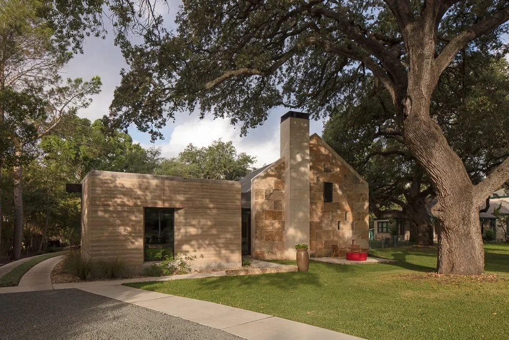 Rammed-earth Hill Country home with stone walls and a standing-seam roof framed by live oaks on a lawn