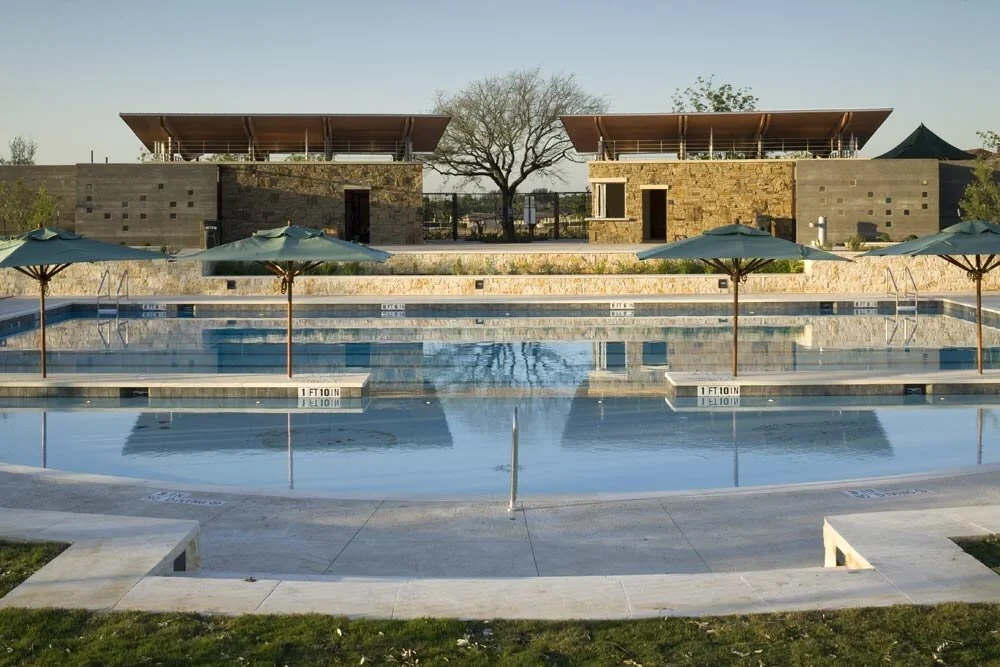 Outdoor lap pool with depth markers below stone-and-timber pavilions and shade umbrellas at sunrise