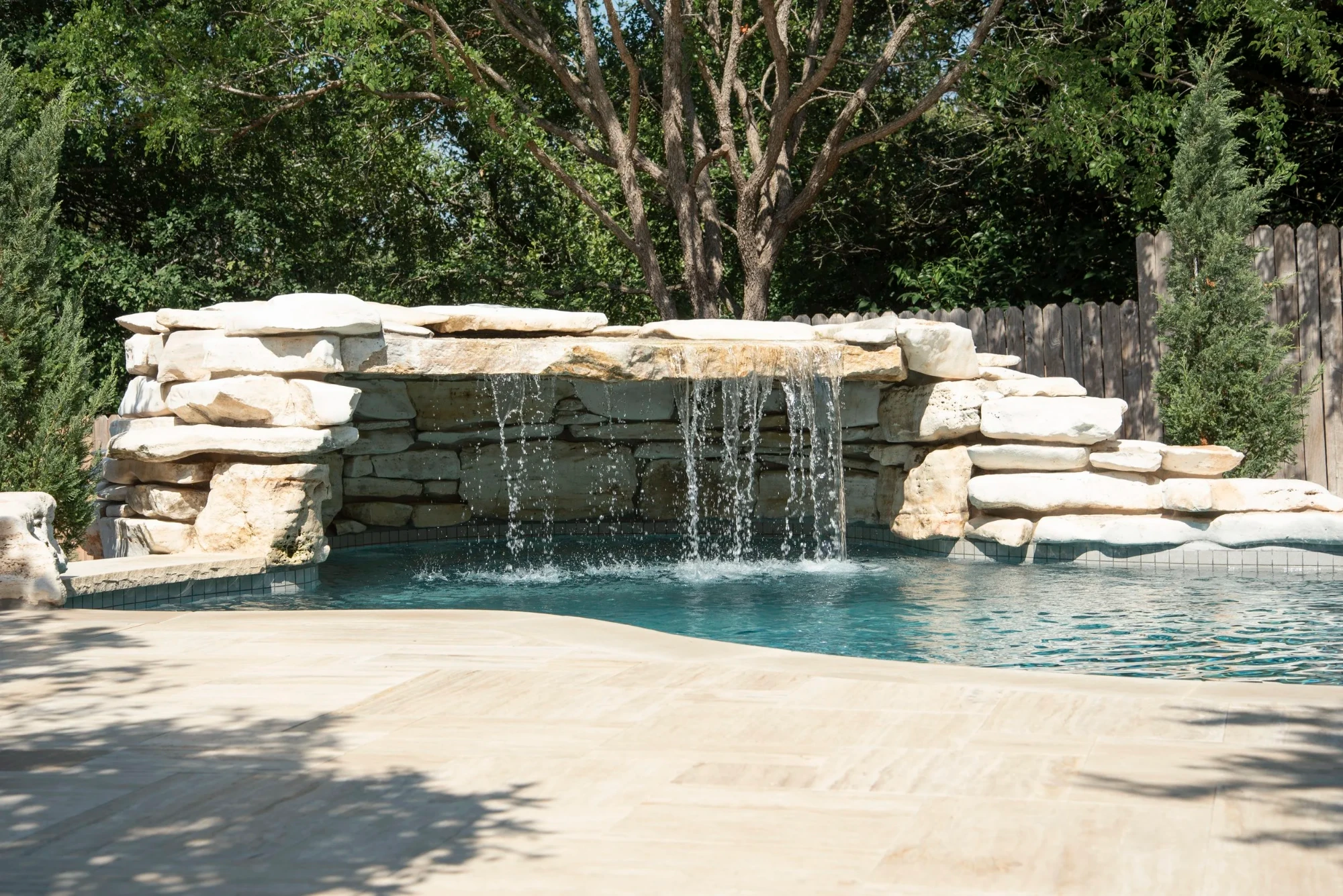 Freeform pool with a stacked-limestone waterfall wall and travertine deck shaded by a tall live oak