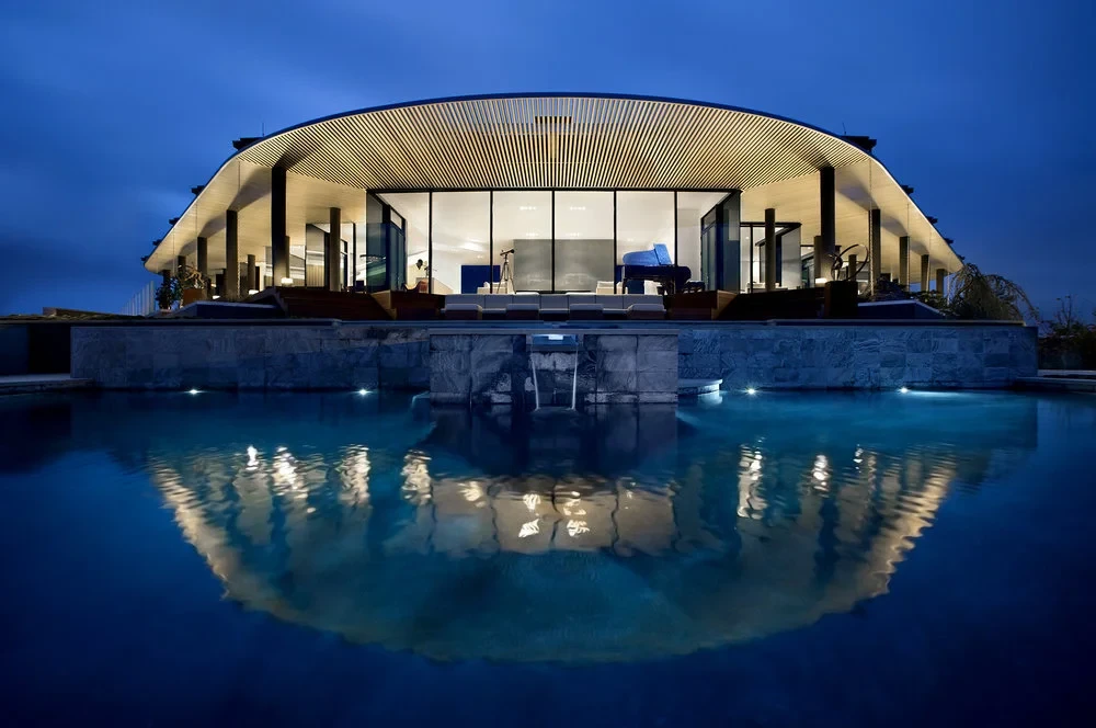 Curved-roof glass home glowing at twilight above a stone-walled pool with a spillway, reflected in still water