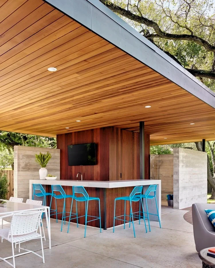 Covered outdoor kitchen with a wood-slat ceiling, stone bar with turquoise stools and a wall-mounted TV