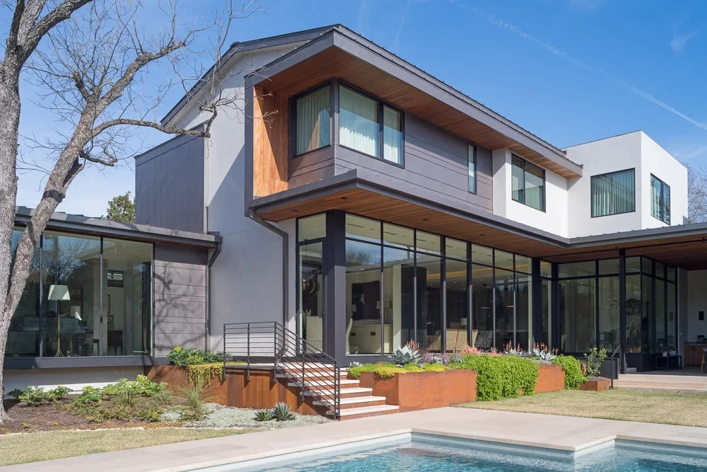 Modern two-story home with wood, stucco and steel siding and walls of glass above a stepped patio and green lawn