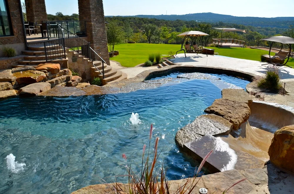 Freeform pool and raised spa with stacked-stone spillways set against a Hill Country home and green valley