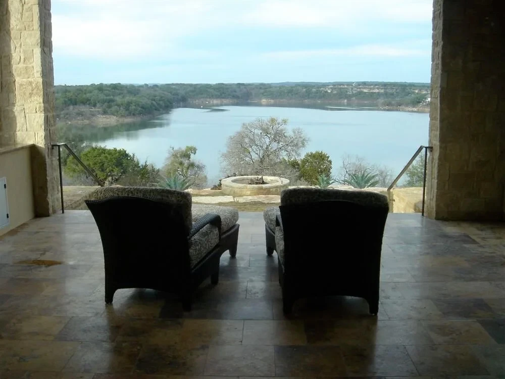 Two wicker chairs on a travertine patio framing a wide lake and wooded hills under stone arches