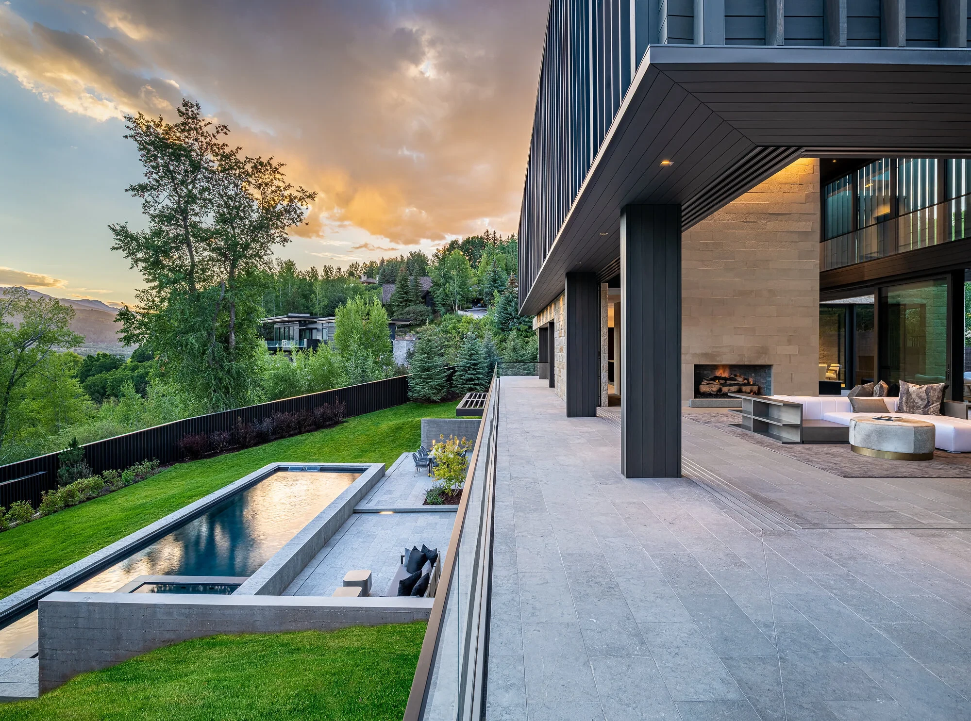 Sunken lap pool and lawn below a modern stone home with a covered patio and fireplace over Aspen hills at dusk