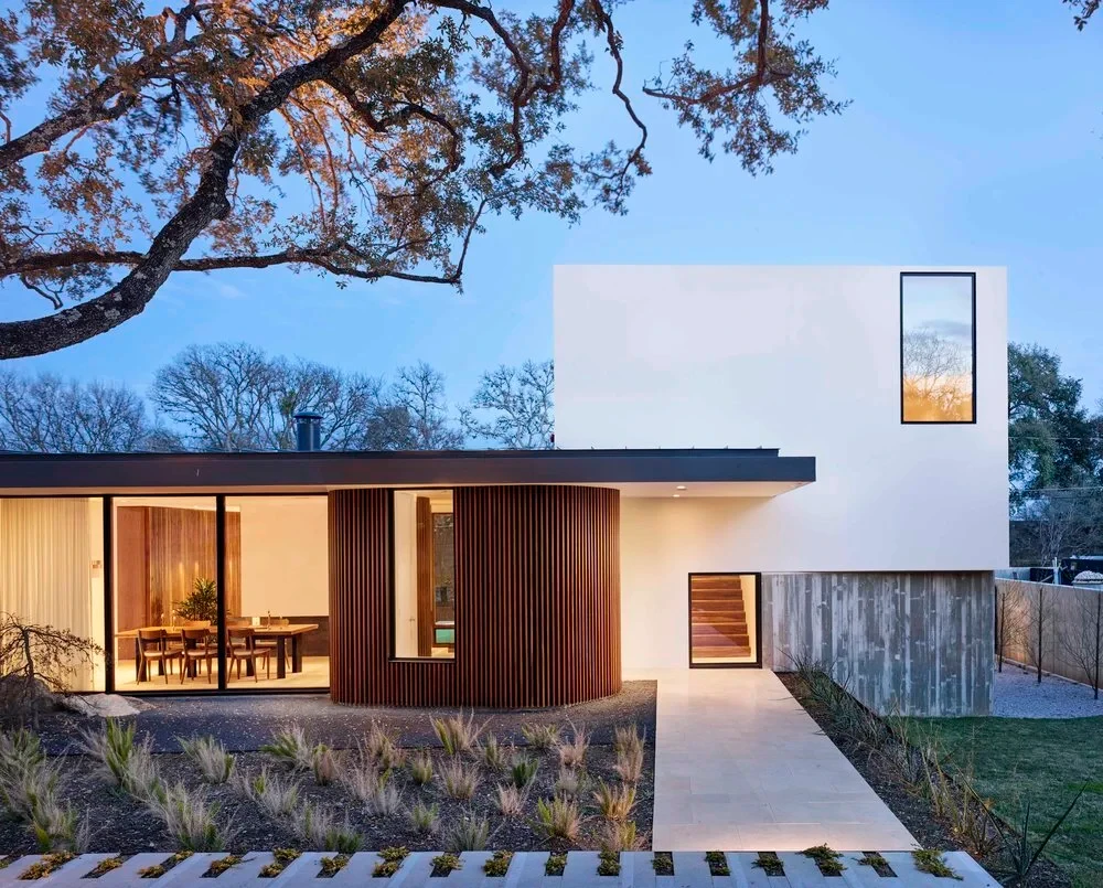 Crisp modern home with white and wood walls and a curved glass corner under oaks at dusk