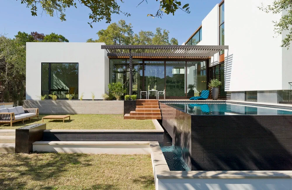 Modern white home with a slat pergola above a dark infinity pool and wood deck on a green lawn