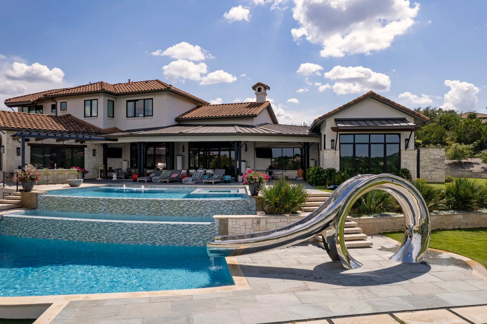 Stainless steel waterslide curling into a tiled pool, with pink blooms and a Lakeway hilltop view