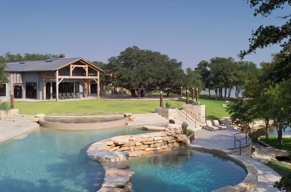 Freeform lagoon pool with stone deck and a spa, framed by a timber lodge and lakeside oaks