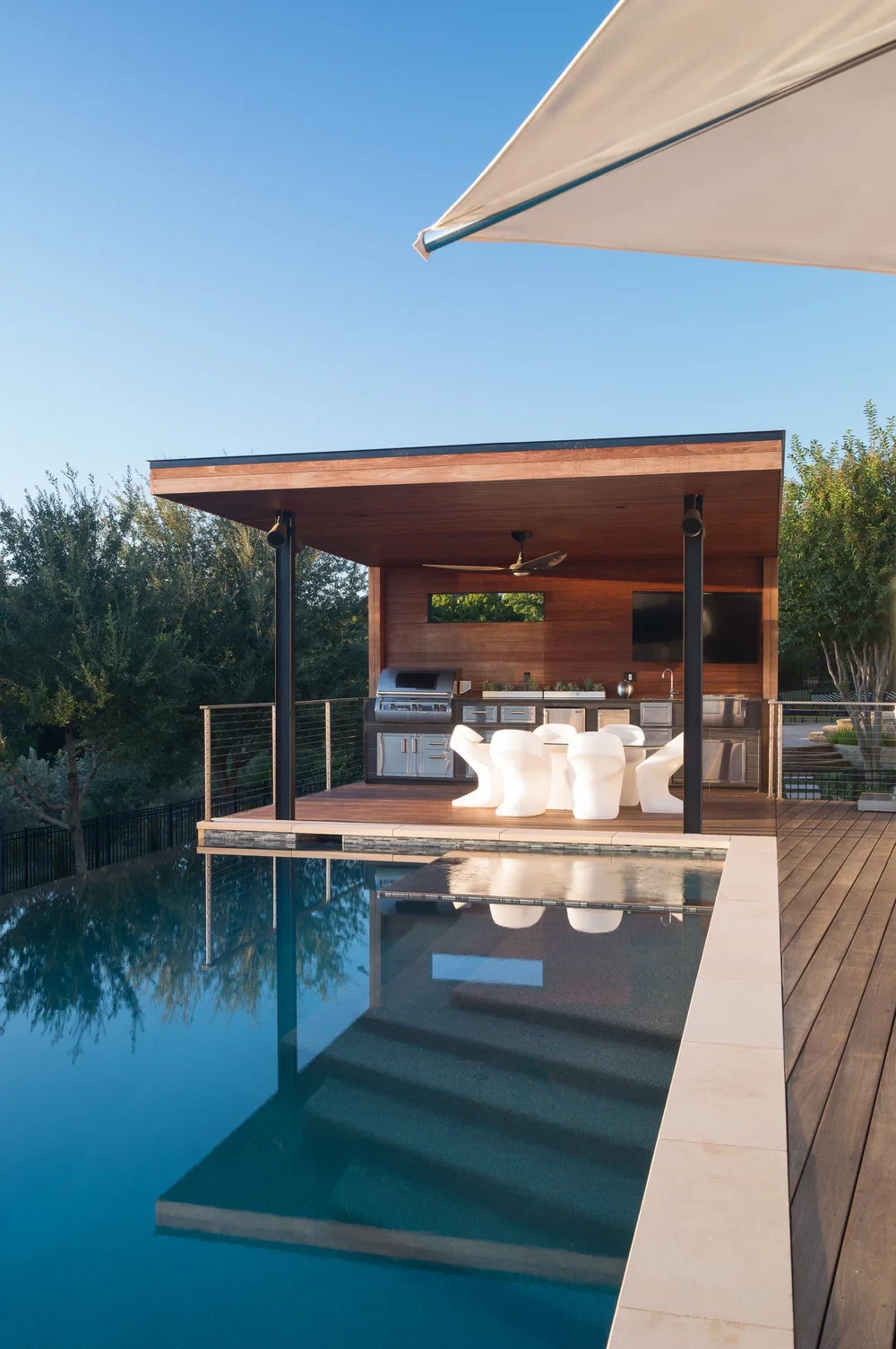 Infinity pool reflecting a wood cabana with outdoor kitchen, over a Hill Country tree line