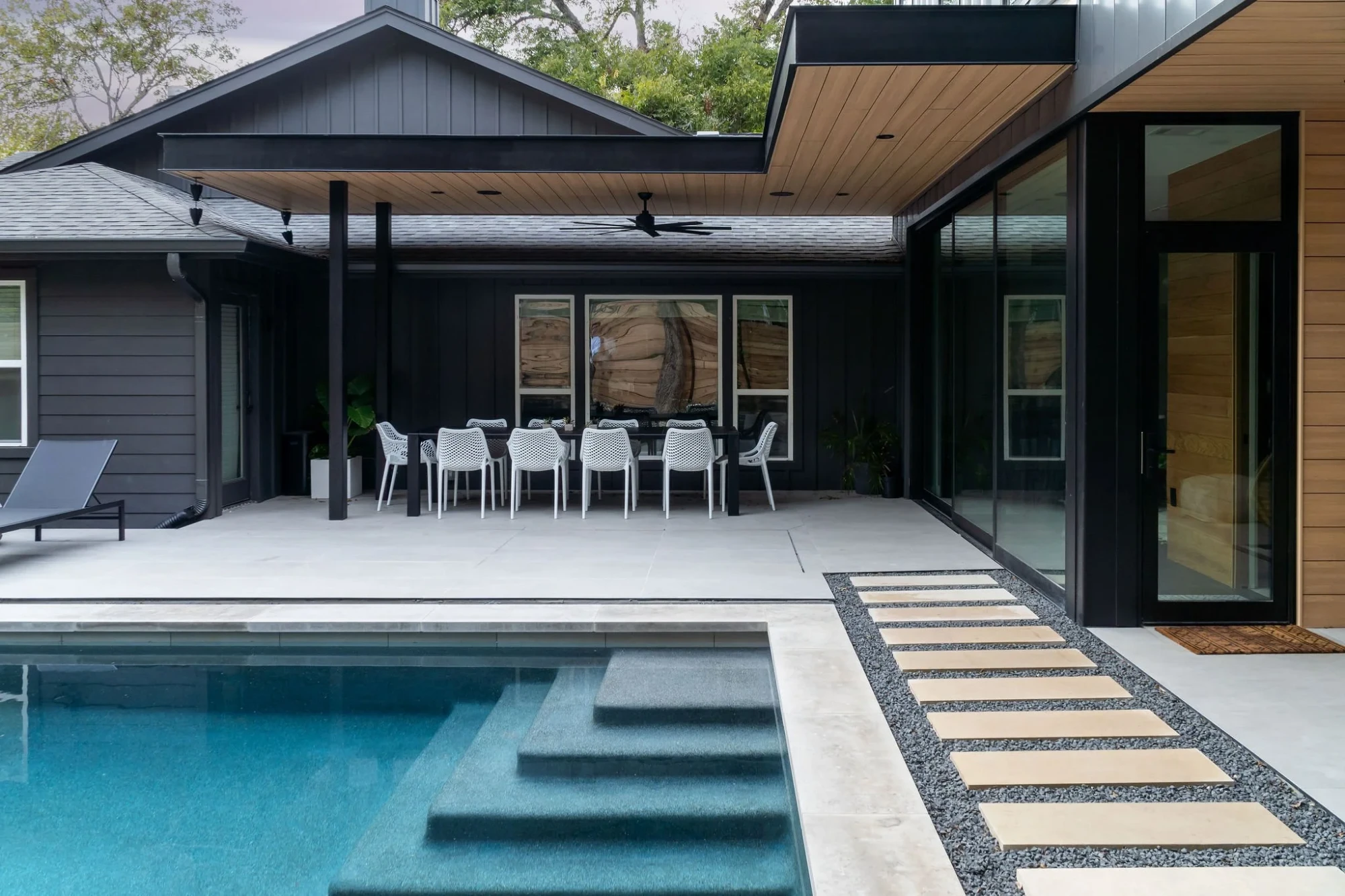 Geometric pool and steps along a stone path beside a black modern home with a covered dining patio