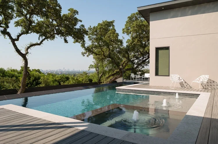 Infinity-edge pool and round spa with bubbling jets on a deck overlooking the Austin skyline through live oaks