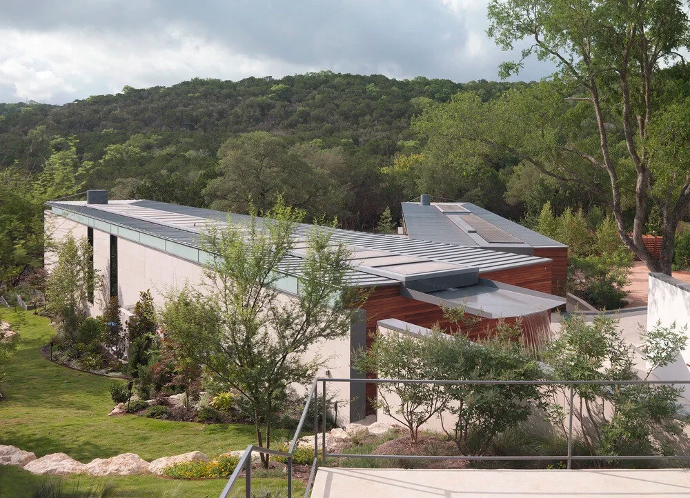 Low modern Hill Country home with metal roof and stone walls set on a green, tree-covered slope