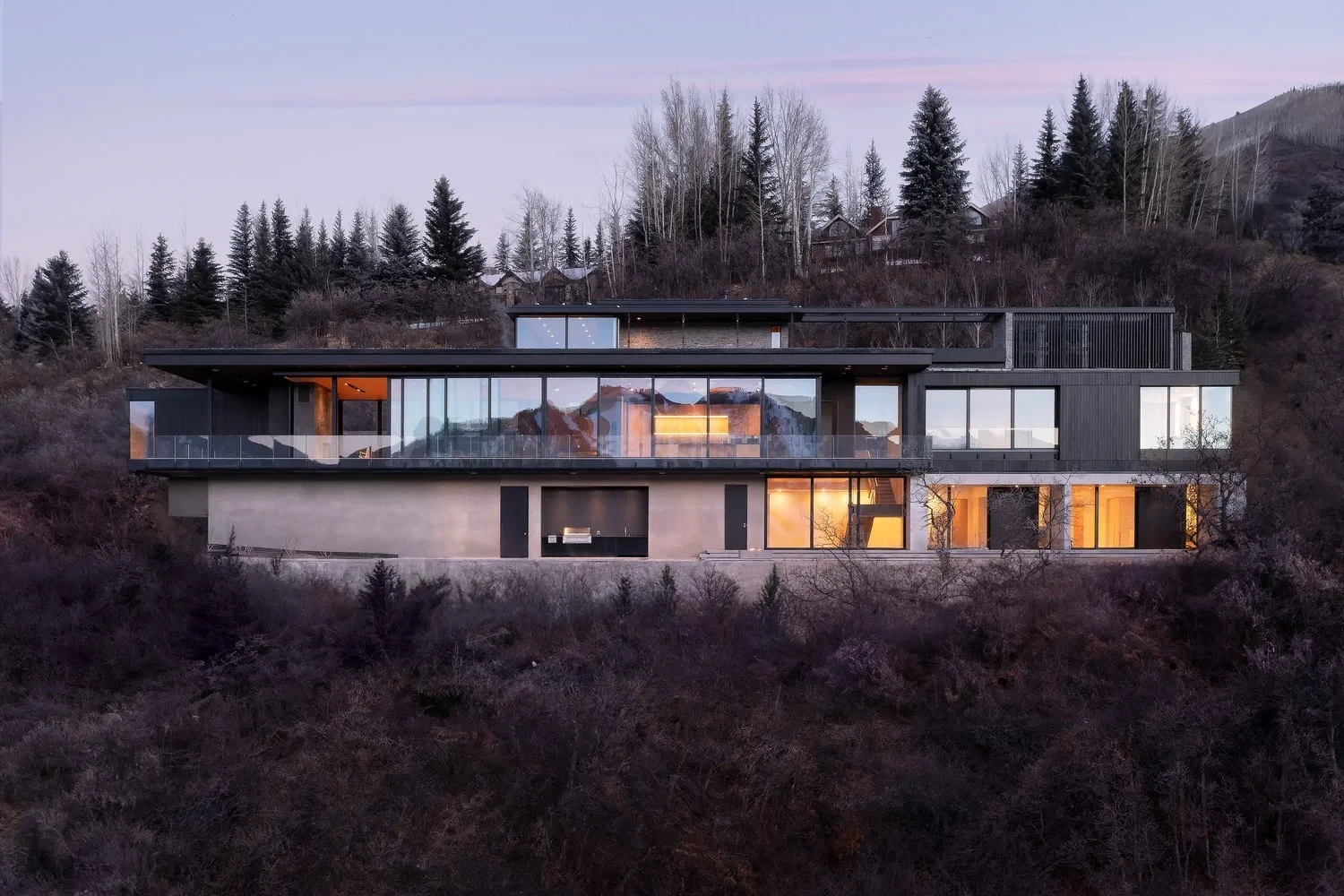Modern glass-and-stone home glowing at dusk on a wooded Aspen hillside below a pink sky