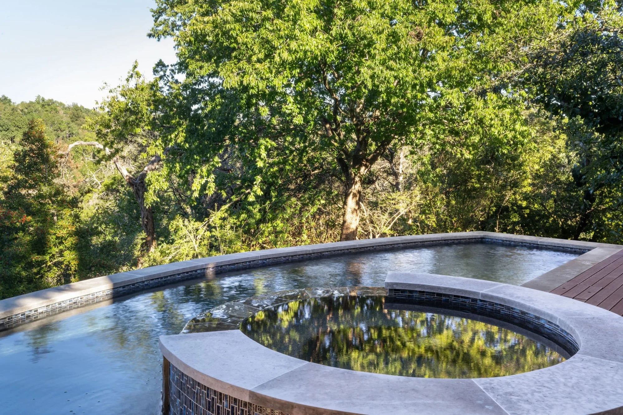 Round spa spilling into an infinity pool with stone coping, overlooking treetops in the Hill Country