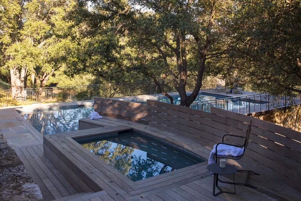 Wood deck wrapping a geometric pool and spa among live oaks on a sloped Hill Country lot