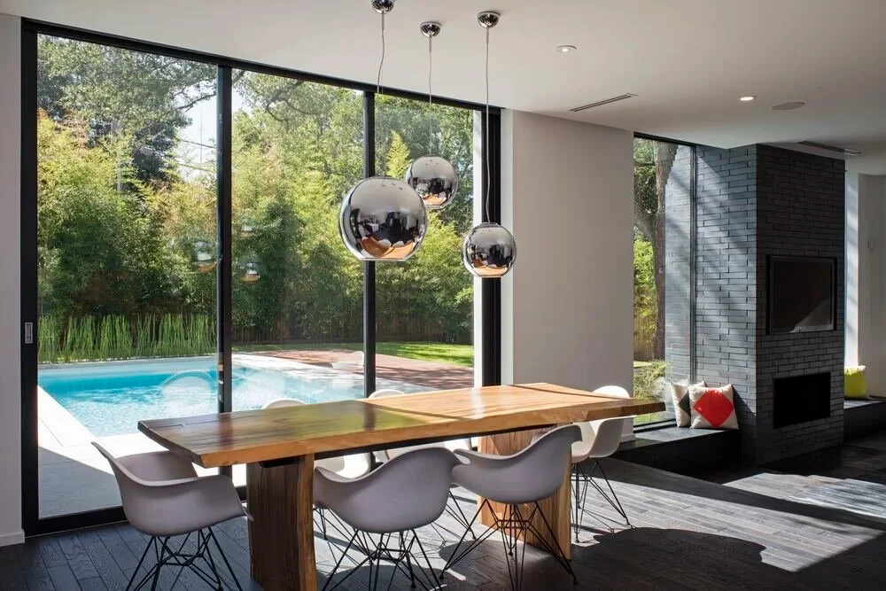 Modern dining room with a live-edge wood table, chrome globe pendants, and floor-to-ceiling glass facing the pool