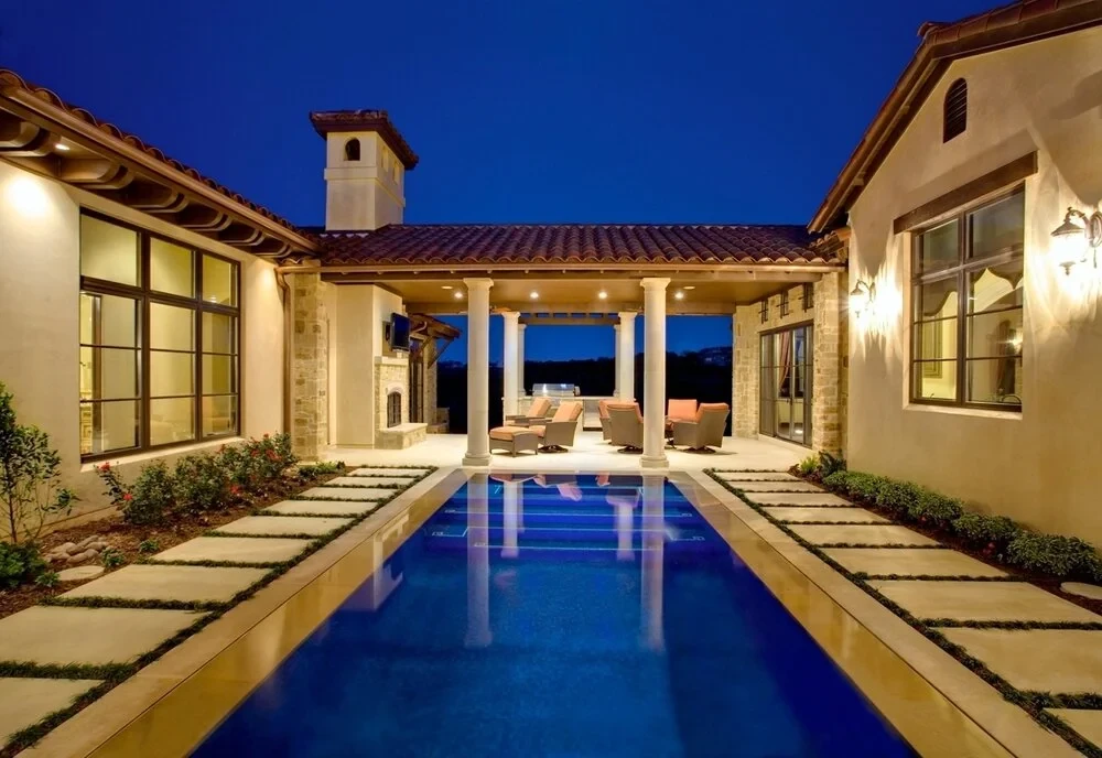 Long geometric lap pool reflecting a covered loggia with stone columns, glowing stone home at dusk
