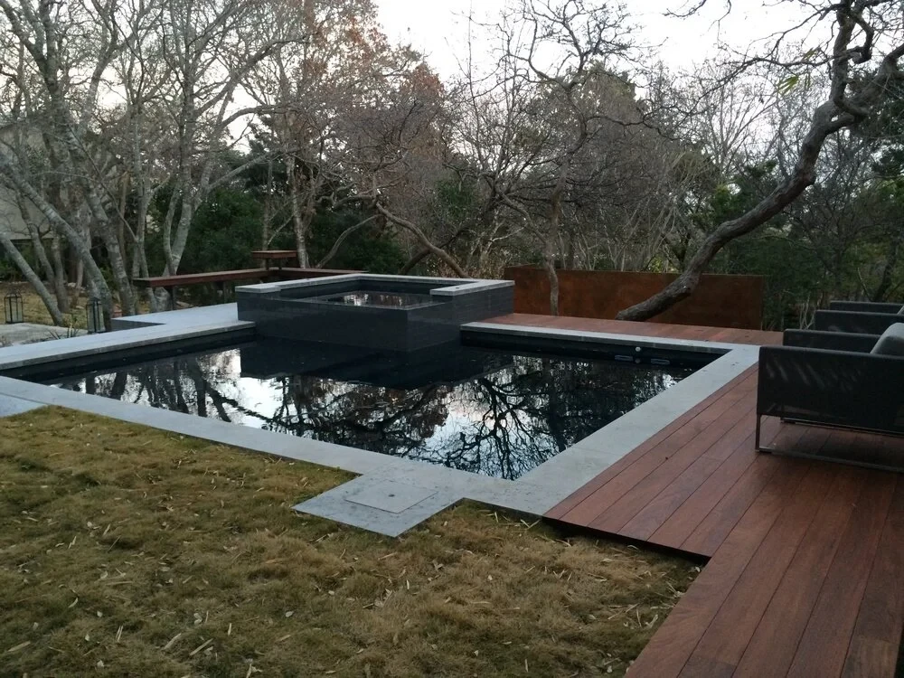 Dark geometric pool with a low spillway and wood deck reflecting bare winter trees in the backyard