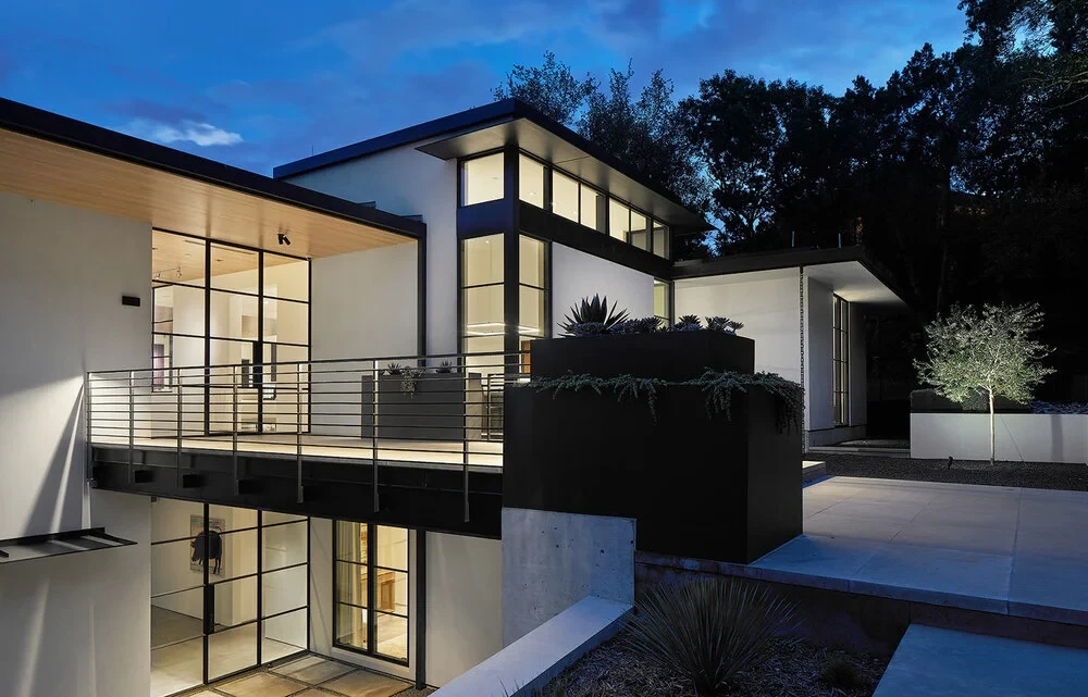 Modern home lit at dusk with glass walls, a steel balcony and a black planter of agave