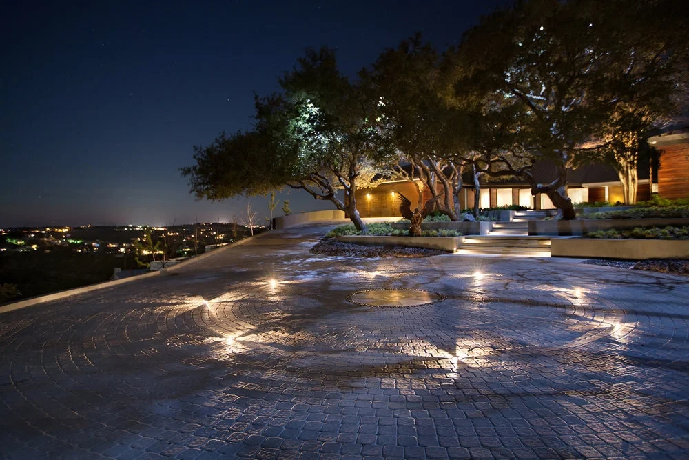 Lit stone deck under live oaks at night, overlooking distant city lights across the Hill Country