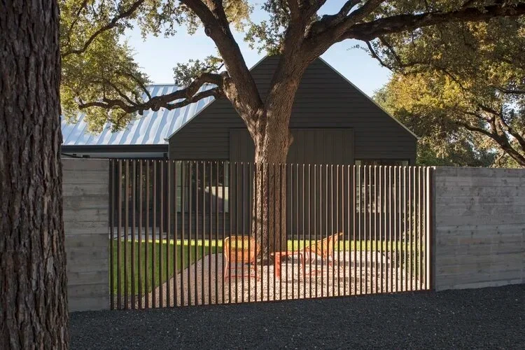 Dark-sided Hill Country home with a metal roof behind a concrete-and-wood slat fence and a live oak