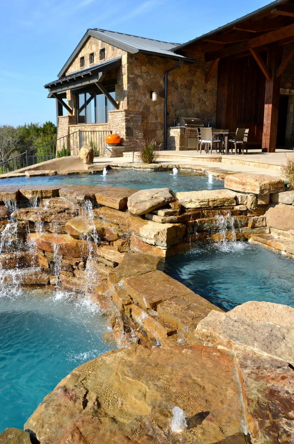 Stacked limestone waterfalls cascading from a raised spa into a freeform pool beside a stone Hill Country home
