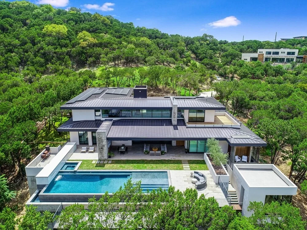Aerial of a modern home with an infinity pool surrounded by green Hill Country woods above Austin