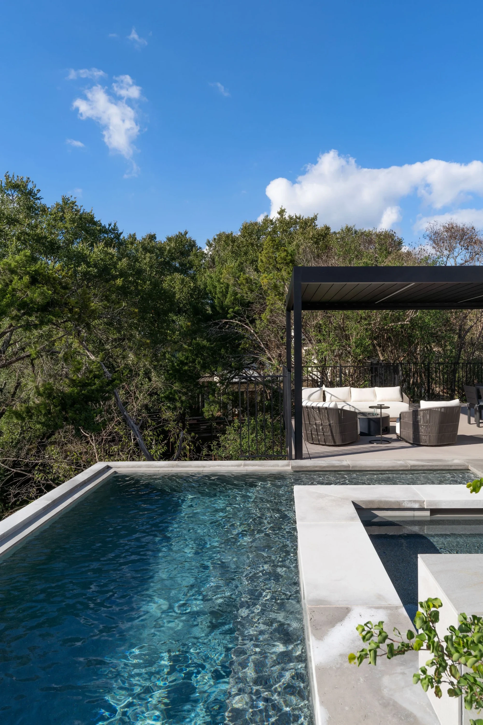 Infinity pool meeting blue sky with a black pergola and seating amid Hill Country treetops