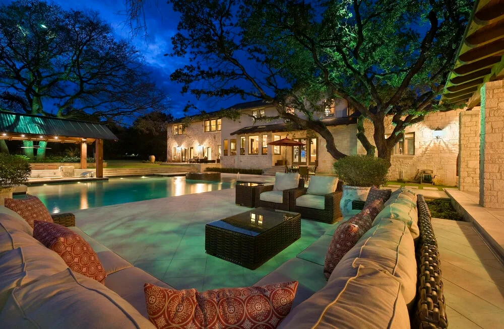 Stone home glowing at dusk above a lit pool with a covered patio and lounge seating in West Lake Hills