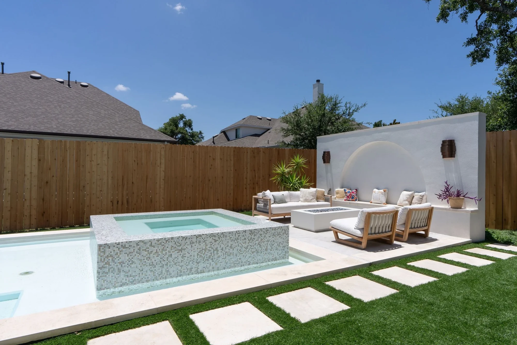 Glass-tile raised spa and geometric pool with a fire table, sectional and stone pavers across the lawn in Austin