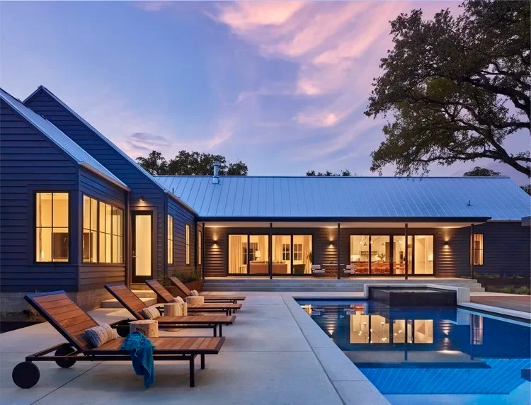 Lake Travis home at dusk with a metal roof, sleek pool and spa, and teak lounge chairs lit warmly indoors