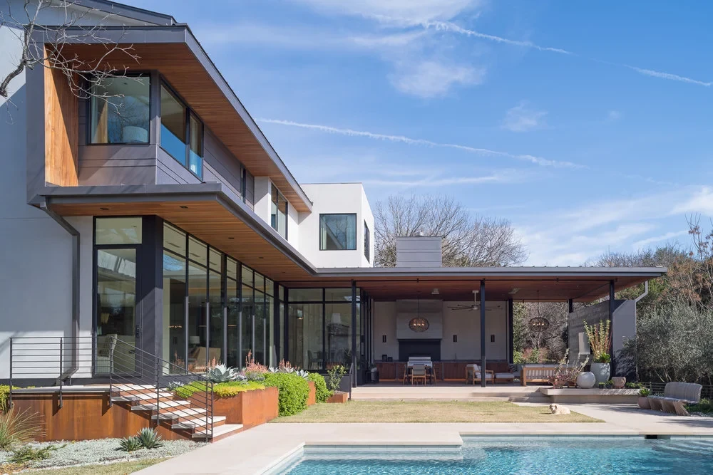 Modern home with a wood-lined cantilevered roof, glass walls and an open-air kitchen above a manicured lawn and pool