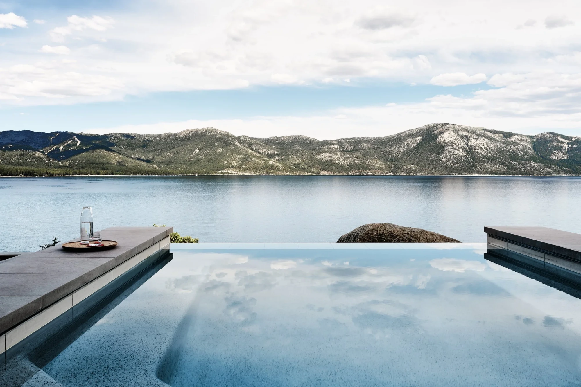 Plunge pool beside a glass home among pines and granite boulders; a tram climbs a rocky hillside