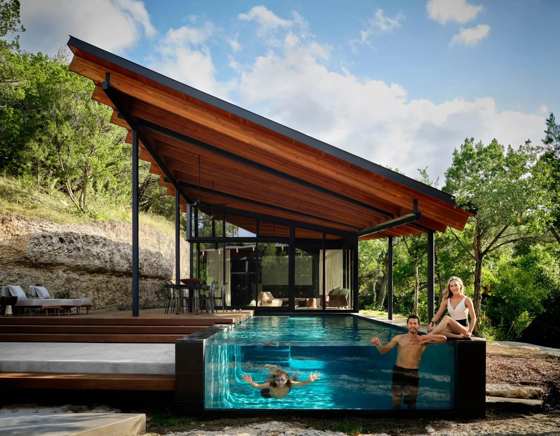Glass-walled geometric pool beside a steel-framed pavilion, with swimmers, a limestone bluff and Hill Country woods
